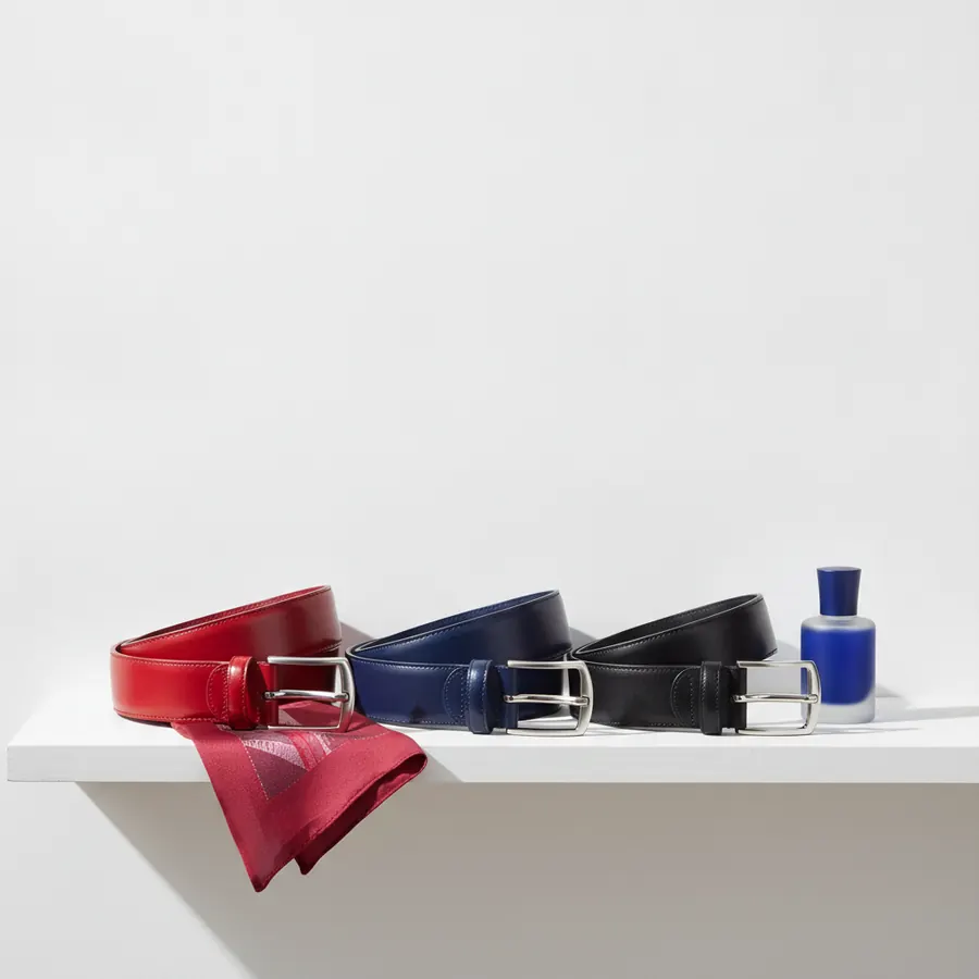 Trois ceintures en cuir rouge, bleu marine et noir avec des boucles argentees posees sur une etagere blanche, accompagnees d'un foulard rouge et d'un flacon bleu et blanc