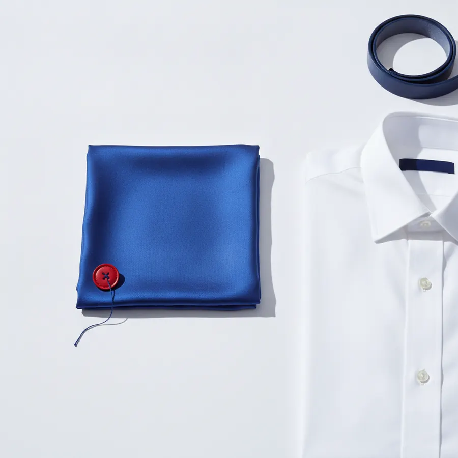 Une pochette carrée bleue satinée avec un bouton rouge cousu dans un coin, une chemise blanche à col classique avec des boutons blancs, et une ceinture enroulée bleu marine, disposés sur un fond blanc