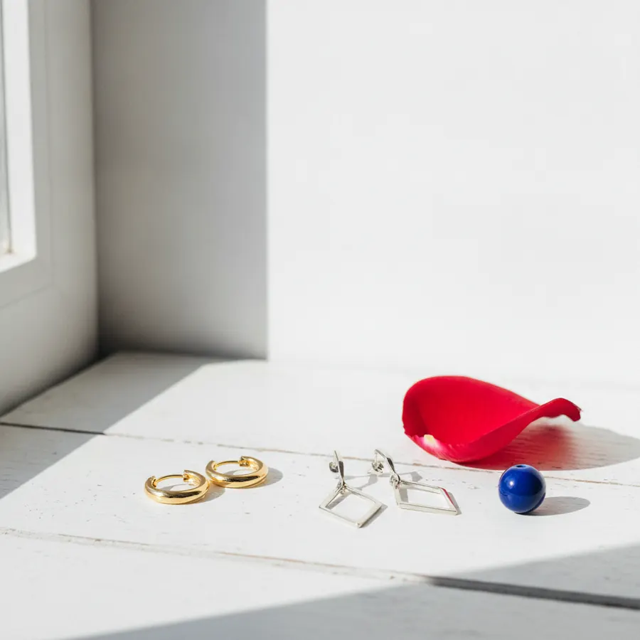 Deux créoles dorées, deux boucles d'oreilles argentées a pendentifs carres, une perle bleue et un petale de fleur rouge, poses sur une surface blanche pres d'un angle de mur blanc