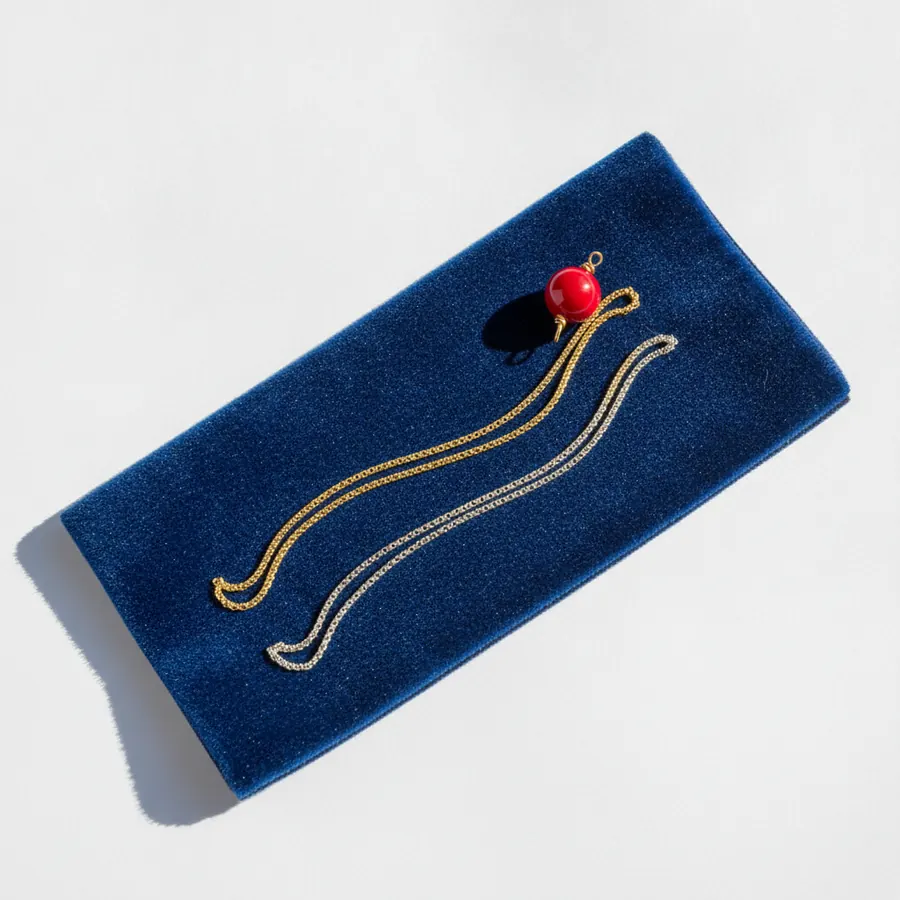 Un rectangle de velours bleu marine sur lequel sont posées deux chaînes fines en métal doré et argenté reliées par un pendentif en forme de boule rouge, sur fond blanc