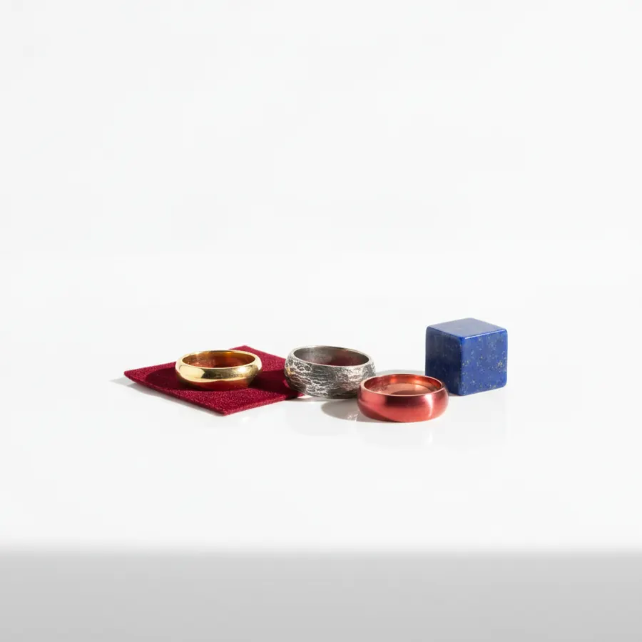 Trois bagues en métal doré, argenté texturé et cuivré rouge posées sur un carré de tissu bordeaux, accompagnées d'un cube bleu marbré, sur fond blanc
