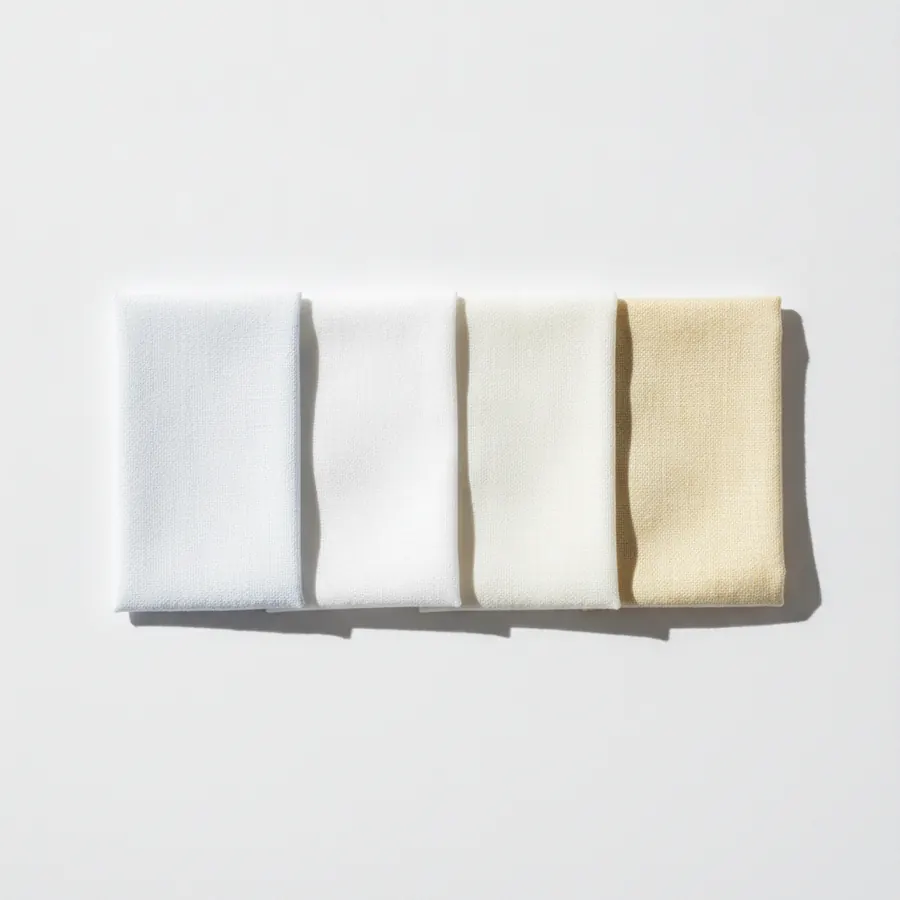 Quatre étoffes blanches posées en ligne sur un fond gris moyen, du blanc optique le plus froid au crème le plus chaud, photographiées en lumière naturelle de côté