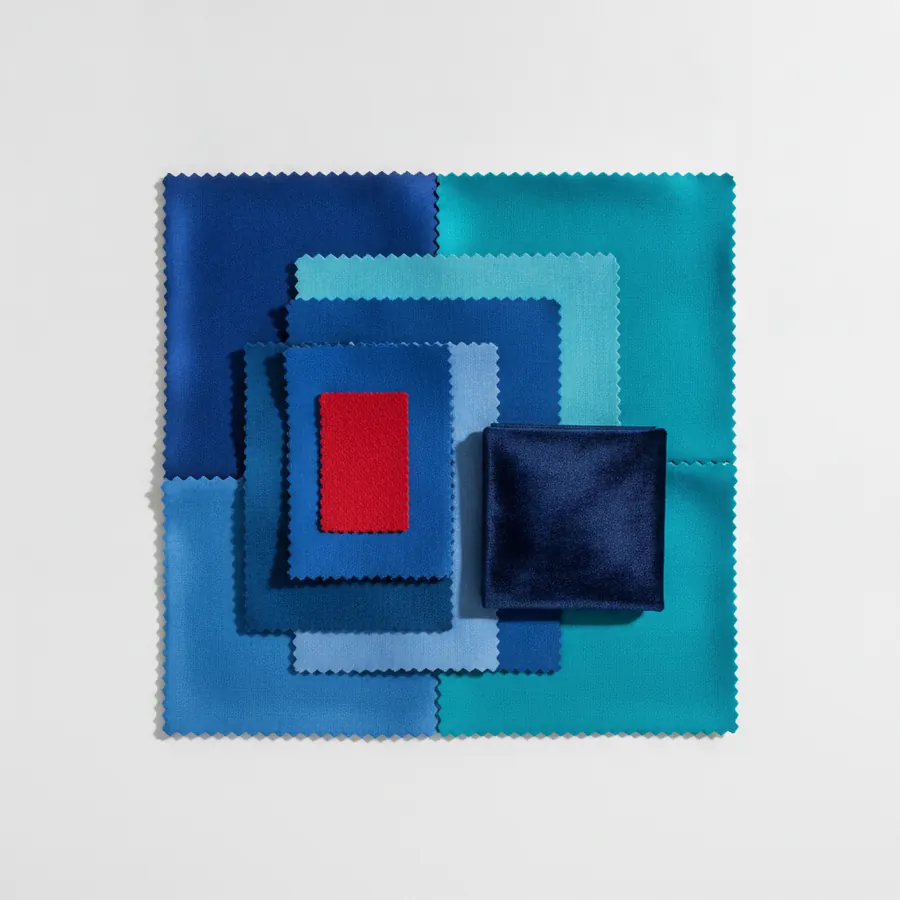 Des echantillons de tissu carres aux bords en zigzag de differentes nuances de bleu et de turquoise sont empiles en decroissant, avec un petit echantillon rouge et une pochette carree bleu marine veloutee poses au centre