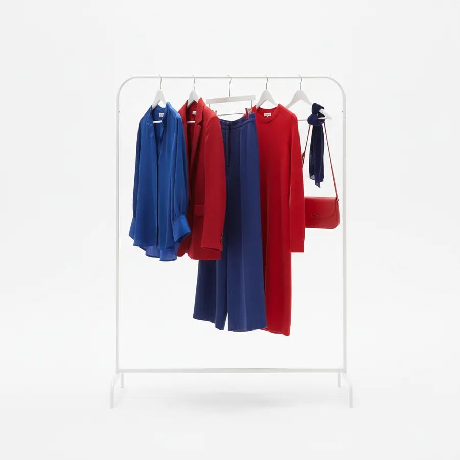Un portant metallique blanc sur lequel sont accroches une chemise bleue, une veste rouge, un pantalon large bleu marine, une robe rouge a manches longues, ainsi qu'un sac a bandouliere rouge et un foulard bleu marine