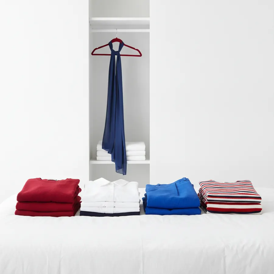Quatre piles de vetements plies en rouge, blanc, bleu et a rayures rouges et bleues posees sur une surface blanche, avec dans l'arriere-plan une niche de placard blanche contenant un foulard bleu marine sur un cintre rouge et des serviettes blanches pliees