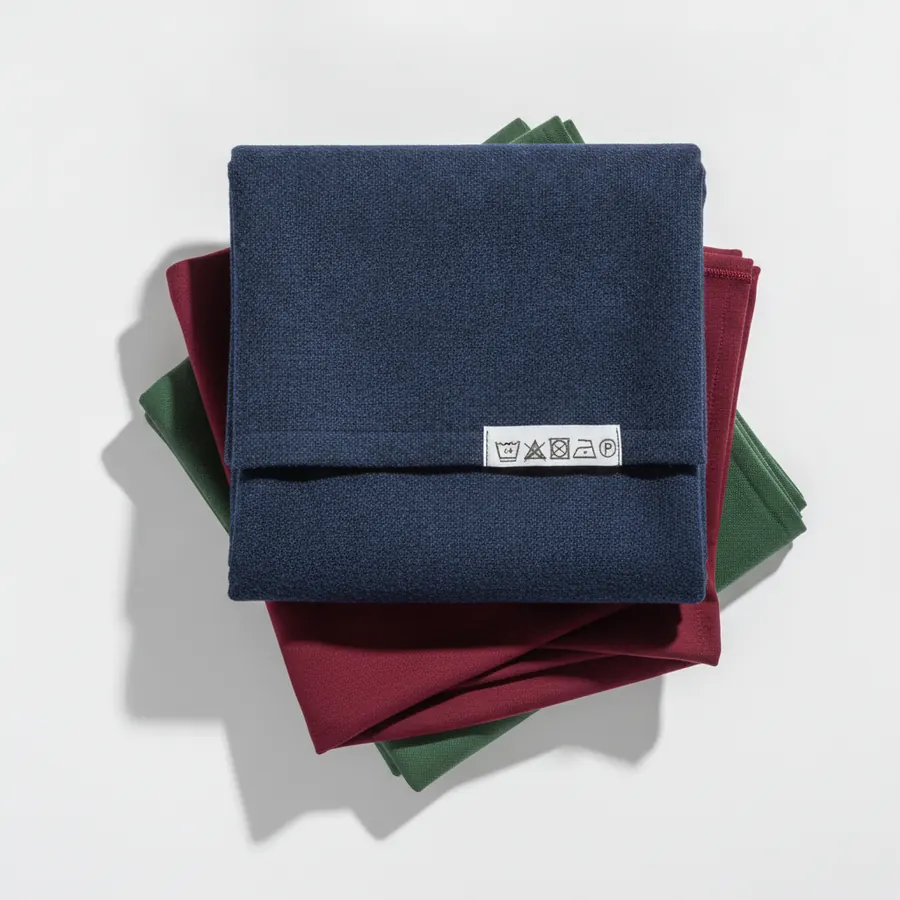 Une pile de trois tissus superposes, un lainage bleu marine, un jersey bordeaux et une toile de coton verte, avec une etiquette de composition visible
