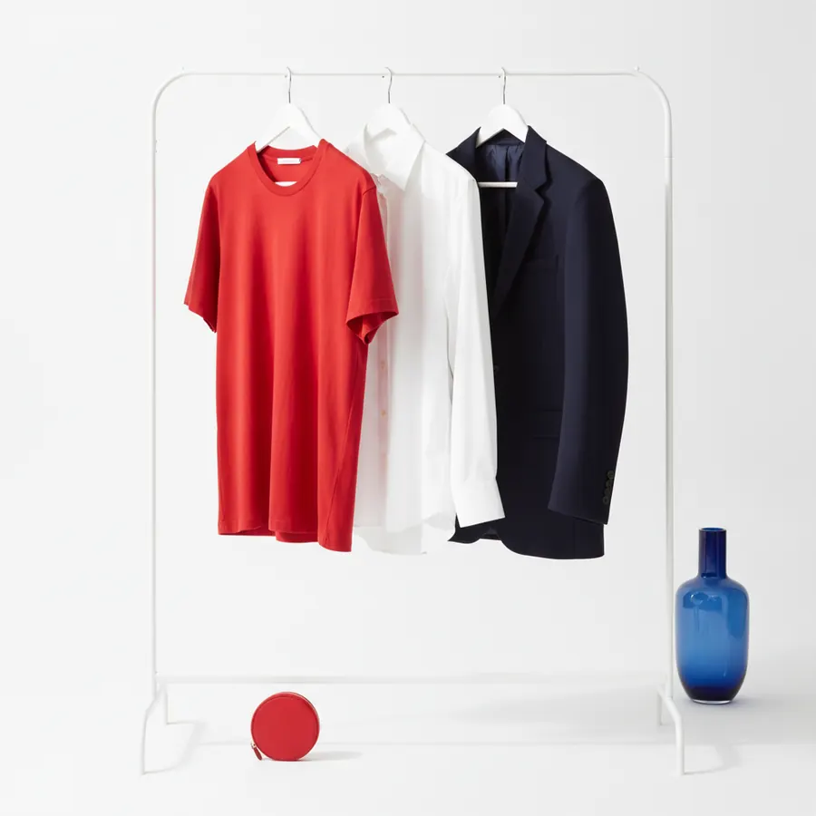 Un portant blanc sur lequel sont accroches un t-shirt rouge, une chemise blanche et une veste de costume bleu marine, avec un petit porte-monnaie rond rouge au sol et un vase en verre bleu a droite