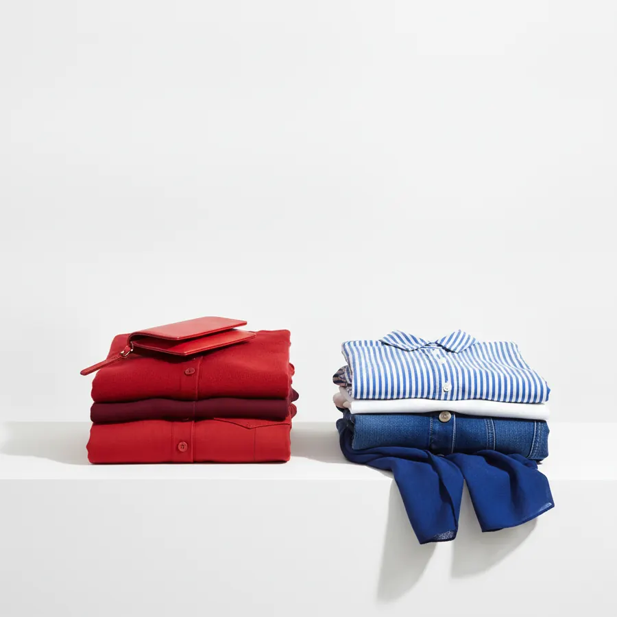 Deux piles de vetements plies poses sur une surface blanche : a gauche, des chemises rouges et bordeaux avec un petit portefeuille rouge en cuir, a droite, une chemise rayee bleu et blanc, un vetement blanc, un jean bleu et un foulard bleu