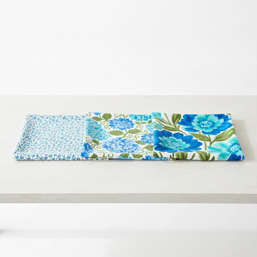 Trois étoffes fleuries posées en ligne sur une table de bois clair, du motif minuscule au très grand motif, dans les mêmes tons bleus et verts, photographiées de dessus