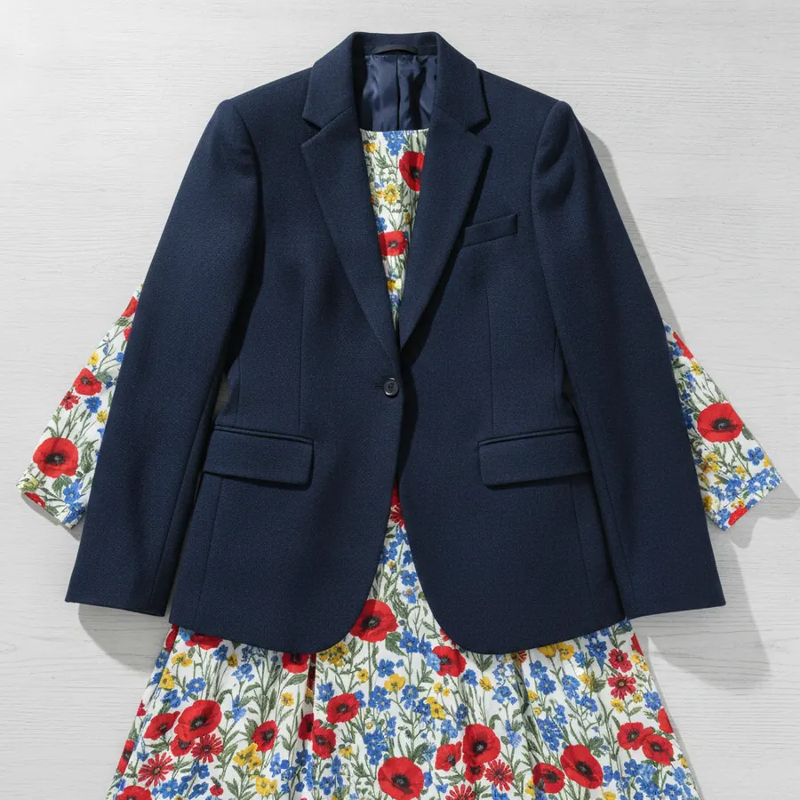 Une veste unie bleu marine posée à plat par-dessus une robe fleurie, sur une table de bois clair, montrant comment la veste casse la surface imprimée