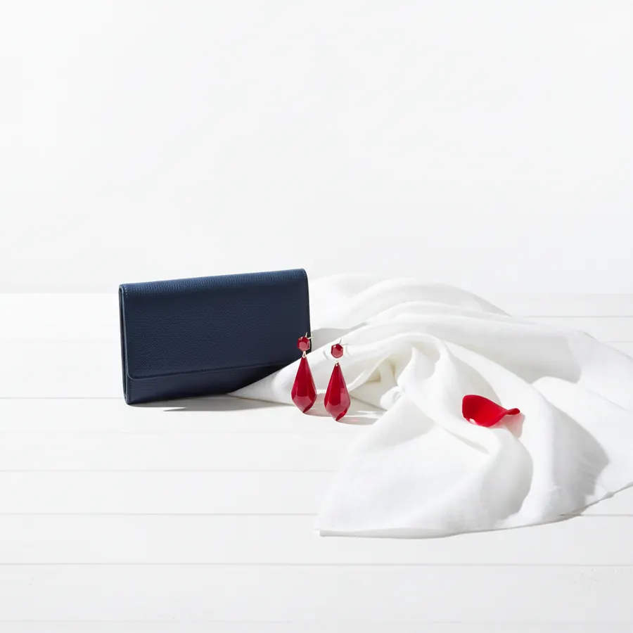 Un portefeuille pochette en cuir bleu marine, une paire de boucles d'oreilles pendantes en pierre rouge et un foulard en soie blanc sont disposes sur une surface blanche