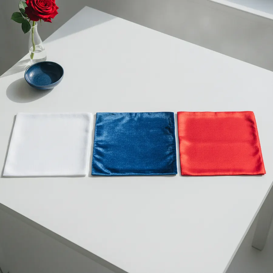 Trois pochettes carrees en tissu satiné blanc, bleu et rouge sont posees cote a cote sur une table blanche, avec un bol bleu et un vase transparent contenant une rose rouge en arriere-plan