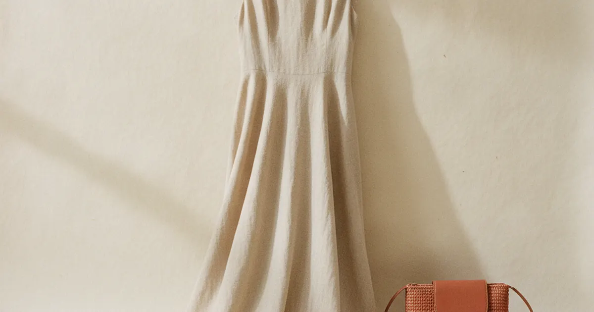 Une robe de ceremonie fluide suspendue a un cintre en bois devant un mur clair, a cote d une pochette et de chaussures plates