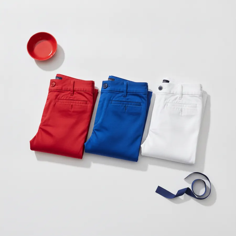Trois pantalons plies cote a cote, l'un rouge, l'un bleu et l'un blanc, disposes sur un fond gris clair, avec un bol rouge en haut a gauche et un ruban bleu marine en bas a droite