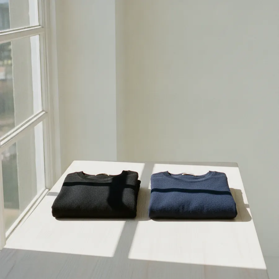 Deux hauts, un noir et un bleu marine, posés à plat sur une table de bois clair près d'une fenêtre, la lumière du jour tombant de côté sur les deux étoffes