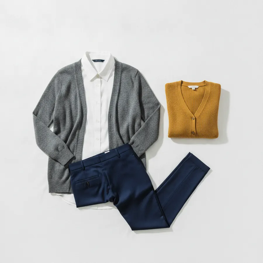 Une tenue disposée à plat sur un lit clair, pantalon marine, chemise blanche et cardigan gris, avec un cardigan supplémentaire plié à côté, vue de dessus