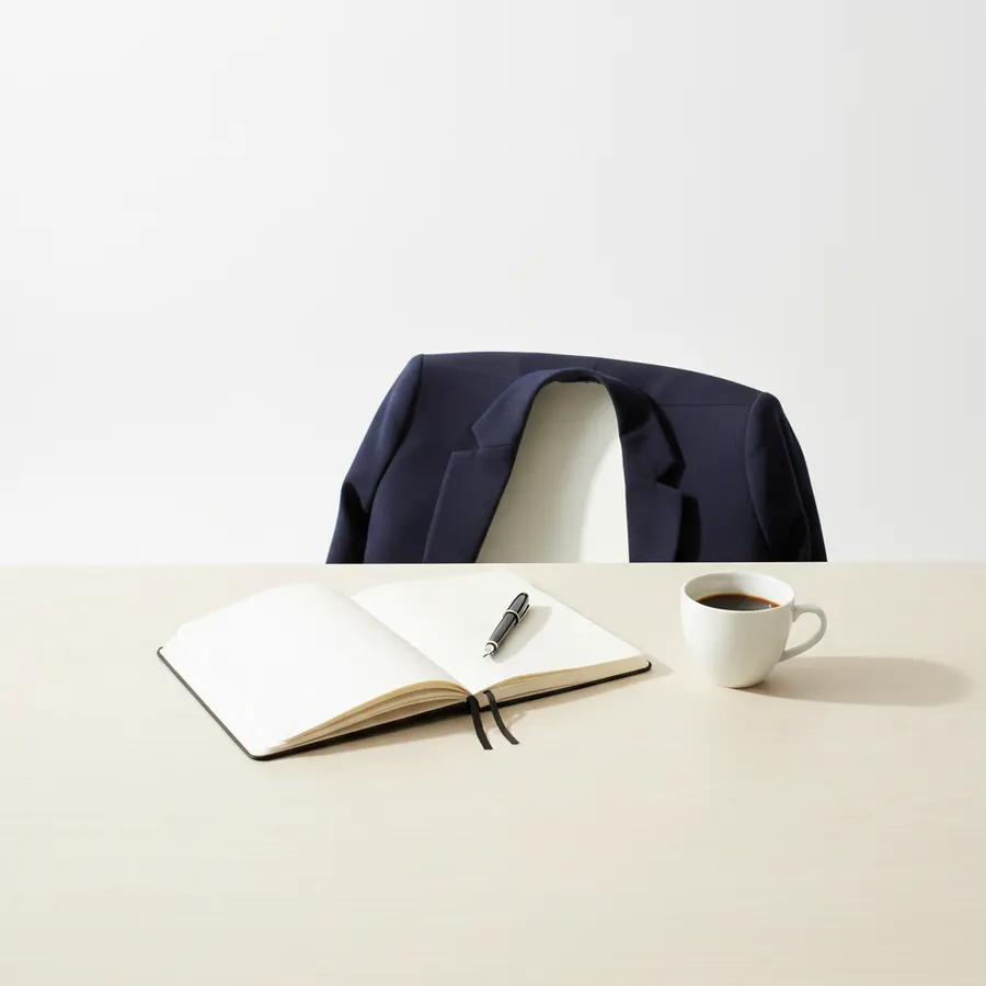 Un carnet ouvert et un stylo posés sur un bureau clair à côté d'une tasse de café, avec une veste marine posée sur le dossier d'une chaise en arrière-plan