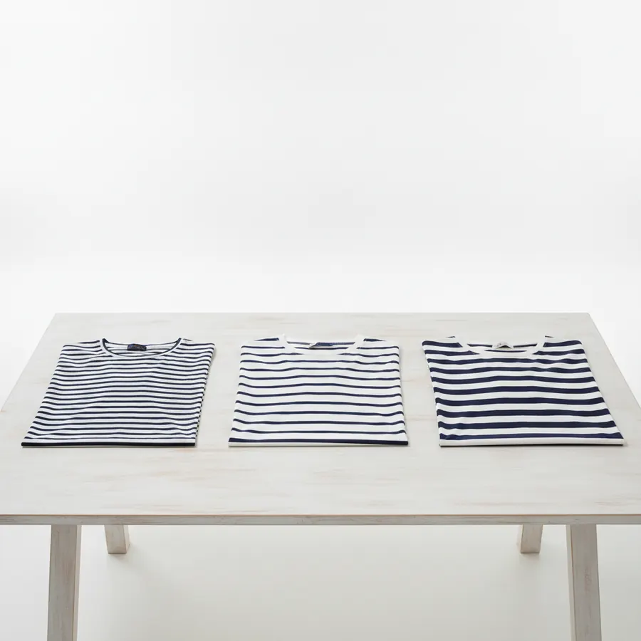 Trois tee-shirts rayés posés à plat sur une table de bois clair, l'un à rayures fines et serrées, l'un à rayures moyennes, l'un à rayures larges, tous en marine et blanc