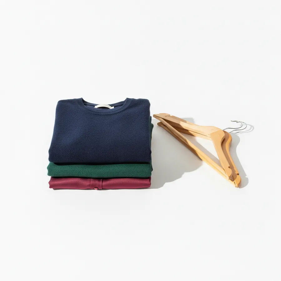 Une pile de vetements plies dans des tons bleu marine, vert foret et bordeaux posee a cote de trois cintres vides sur fond blanc