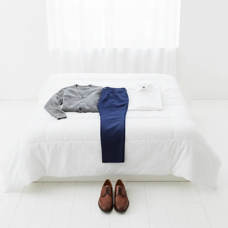 Une tenue préparée sur un lit à la couette blanche, un cardigan gris à gauche, un pantalon bleu marine au centre et une chemise blanche pliée à droite, avec une paire de derbies en cuir cognac posée sur le parquet clair au pied du lit