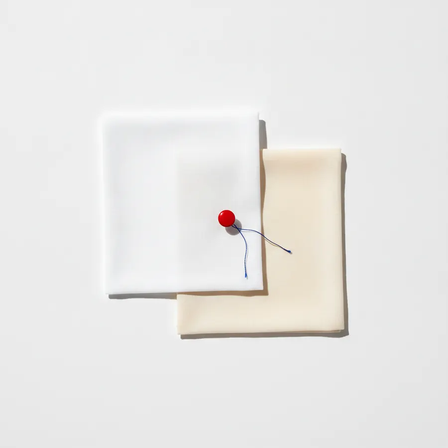 Deux carre de tissu plies superposes sur un fond blanc, l'un blanc et l'autre beige creme, maintenus par une epingle a tete ronde rouge avec un fil bleu