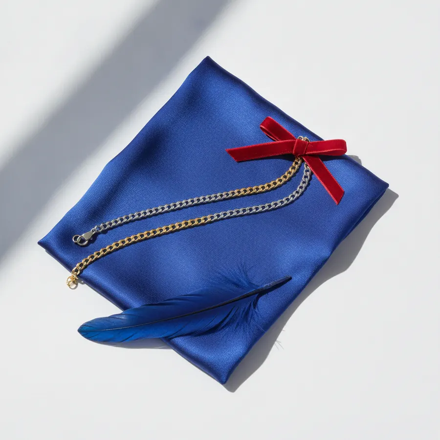 Une pochette en tissu satin bleu cobalt posee sur un fond blanc, sur laquelle reposent deux chaines fines l'une argentee l'autre doree, un petit noeud en velours rouge et une plume bleue et noire