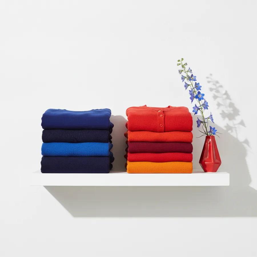 Une etagere blanche fixee au mur sur laquelle sont posees deux piles de pulls plies, l'une dans les tons bleu marine, noir et bleu electrique, l'autre dans les tons rouge, bordeaux et orange, avec un vase rouge geometrique contenant des fleurs bleues