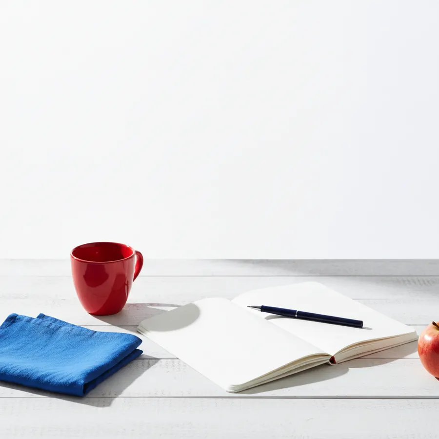 Sur une surface en bois blanc, une tasse rouge, un carnet ouvert aux pages blanches avec un stylo bleu marine pose dessus, un tissu plie bleu et une pomme rouge