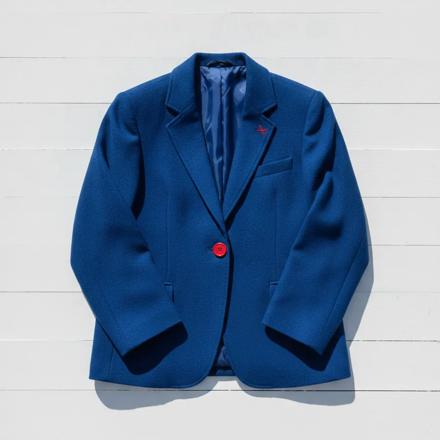 Un blazer bleu roi a bouton unique rouge et a petit motif brode rouge en croix sur le revers, pose a plat sur un fond de planches blanches horizontales