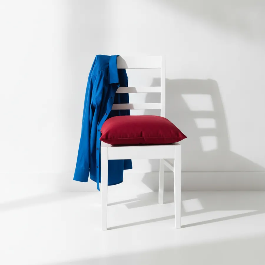 Une chaise blanche avec un coussin rouge posé sur l'assise et une chemise bleue accrochée au dossier, sur un fond blanc