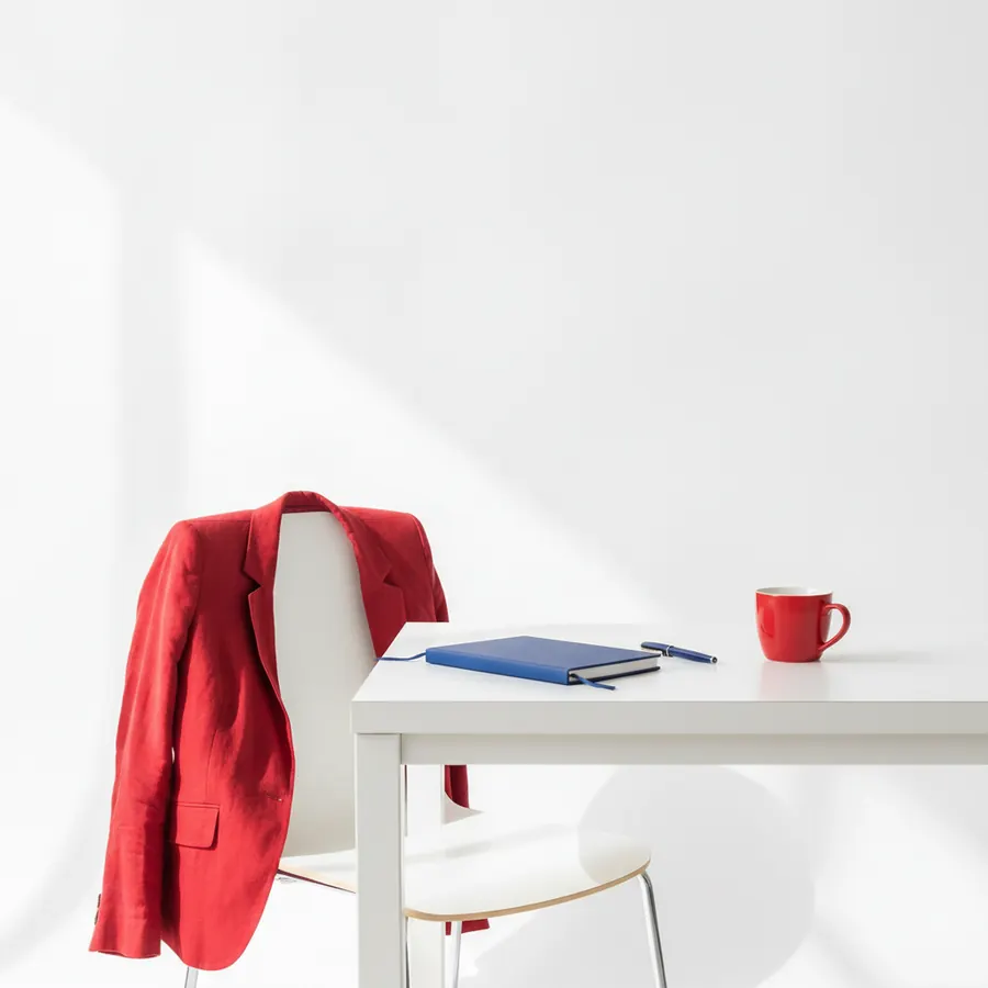 Une veste rouge posée sur le dossier d'une chaise blanche devant un bureau blanc, sur lequel sont poses un carnet bleu, un stylo bleu et une tasse rouge